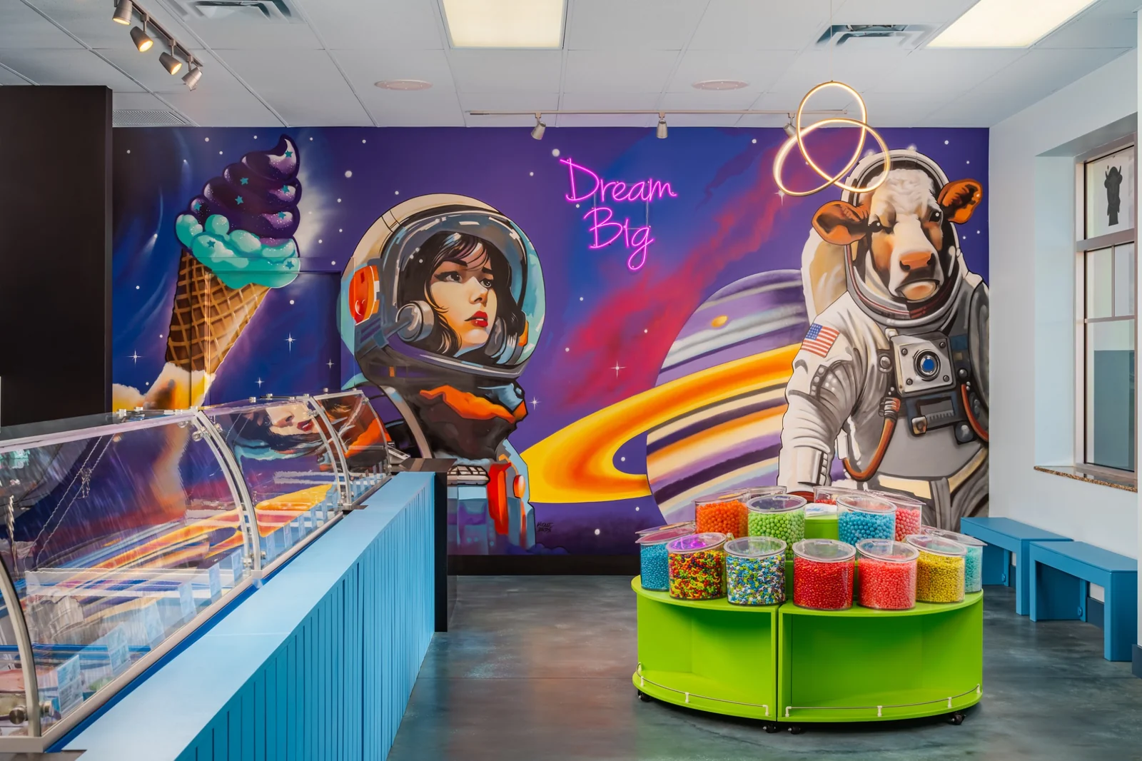 A colorful space mural with an astronaut girl, an astronaut cow, and a giant ice cream cone behind the ice cream case.