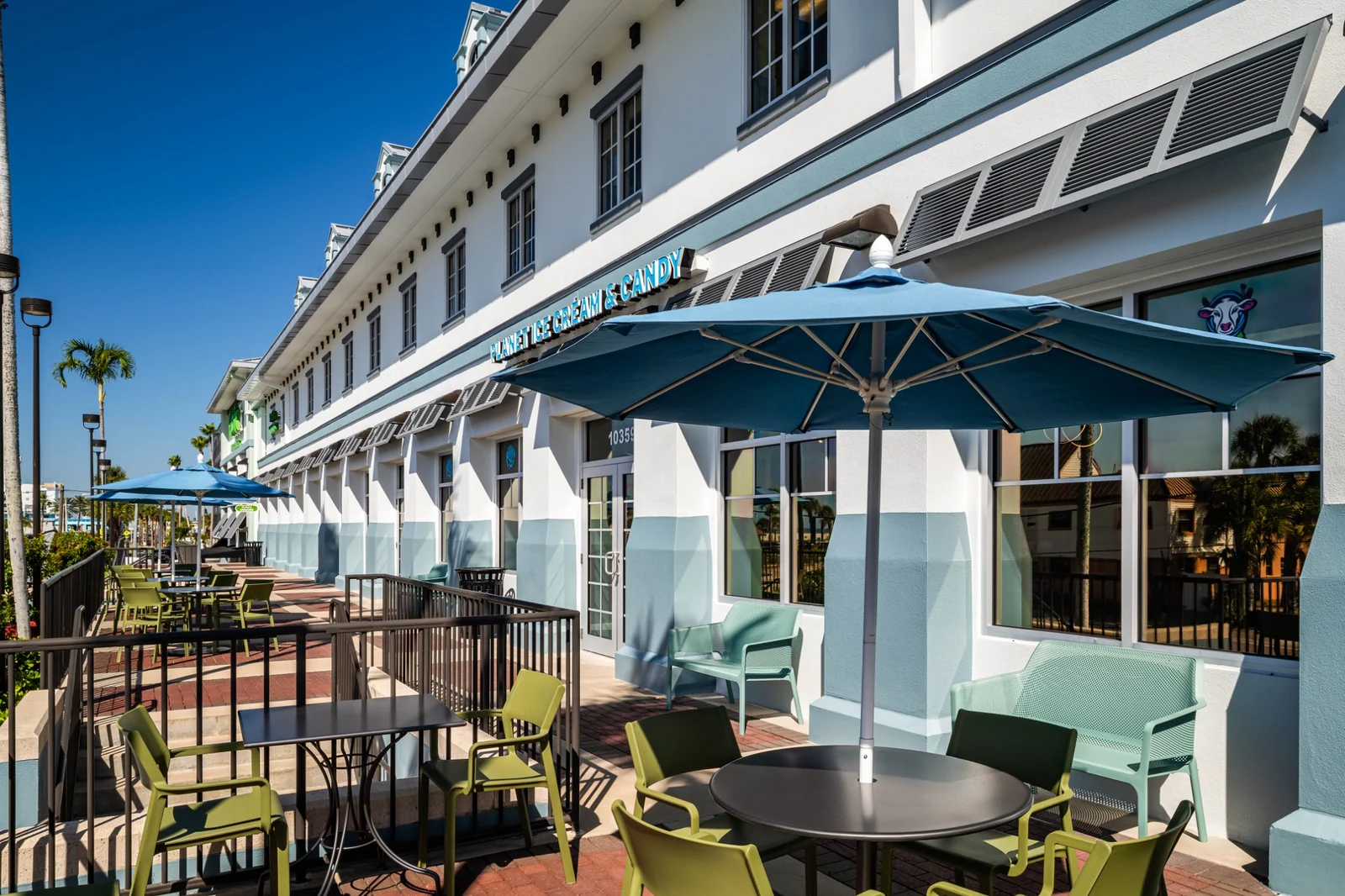 A wide view of patio tables, mint-green chairs, navy umbrellas, and Planet’s storefront along the outdoor walkway.
