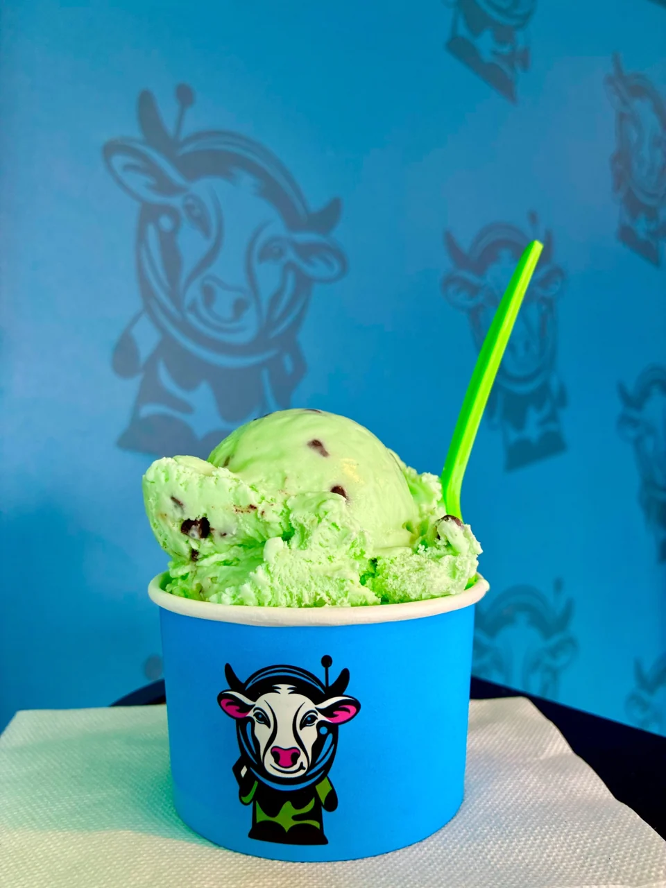 Green ice cream with dark pieces in a blue astronaut-cow cup, with a green spoon against a blue wall decorated with the cow mascot.