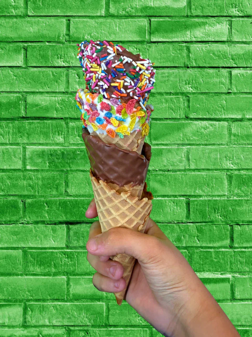 A hand holds a sprinkle-coated waffle cone with ice cream against a green brick wall.