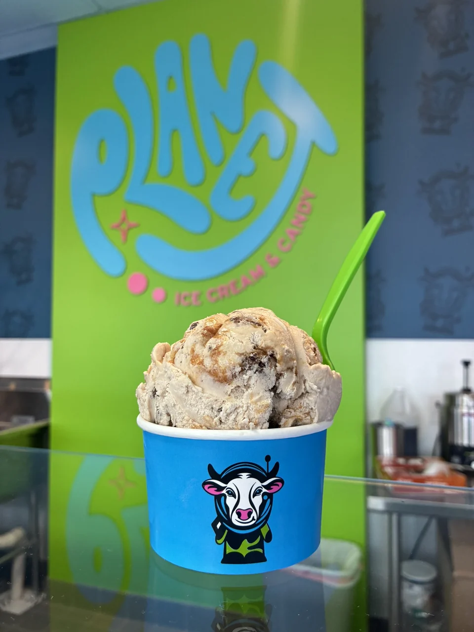 A scoop of cream-colored ice cream with dark pieces in a blue astronaut-cow cup, with a green spoon and the Planet logo behind it.
