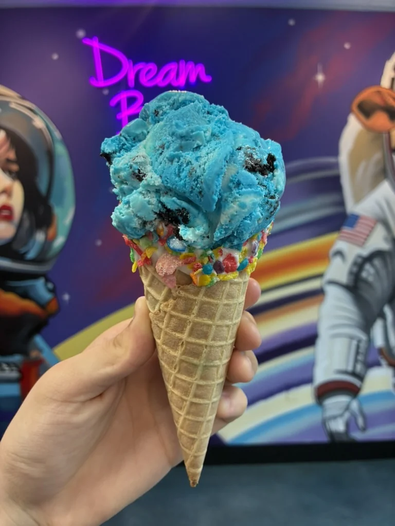 A hand holds a waffle cone with a bright blue scoop and a colorful cereal-coated rim in front of the space mural.