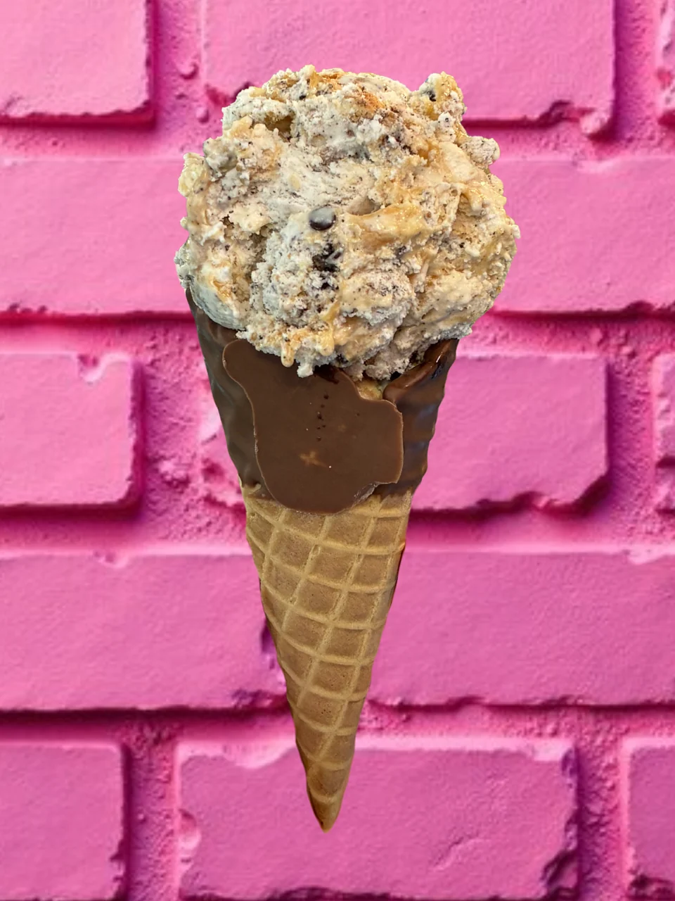 A cream-colored scoop with dark and golden pieces in a waffle cone with a chocolate-dipped rim, against a pink brick wall.