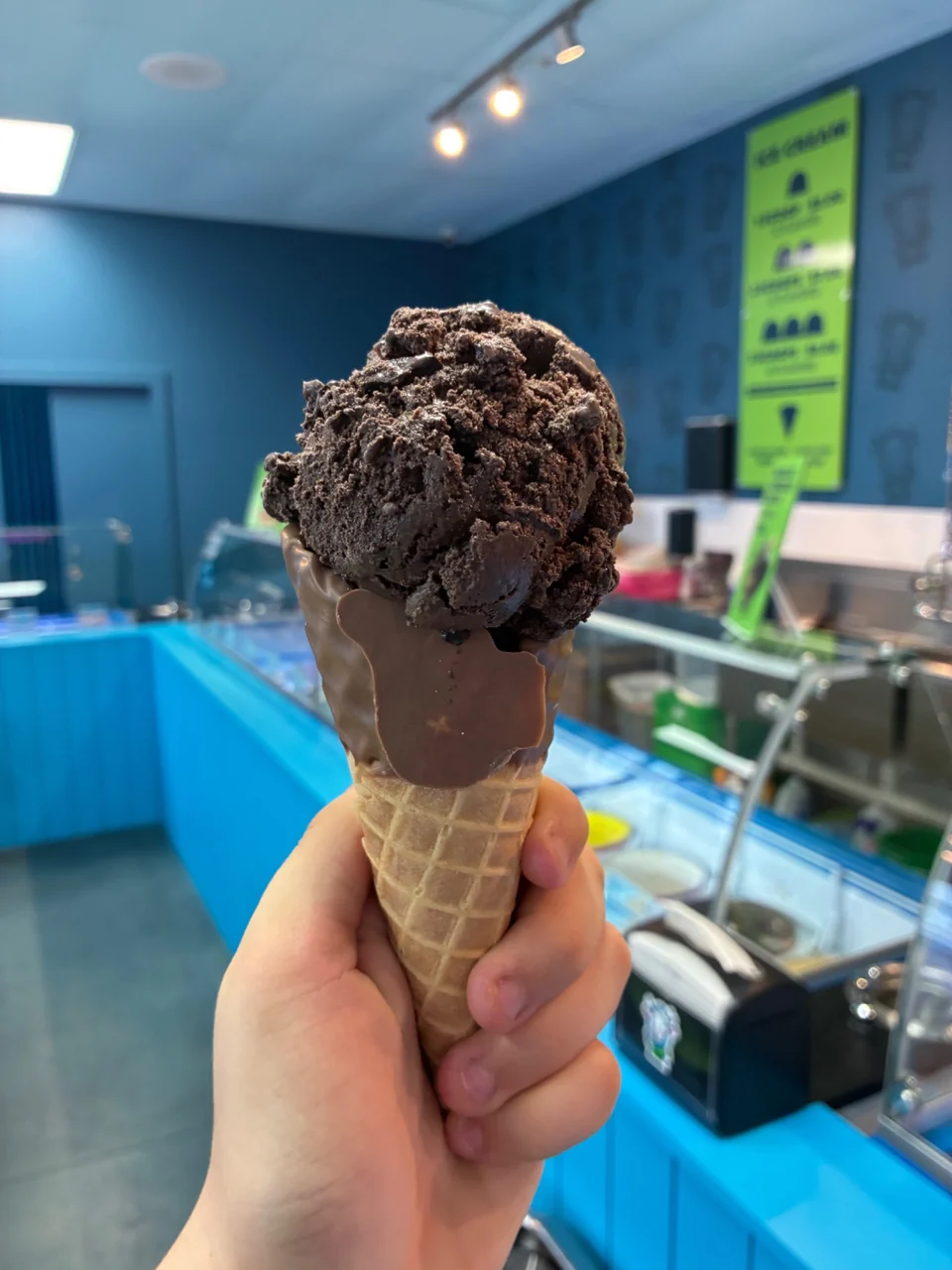 A hand holds a chocolate-coated waffle cone topped with a dark chocolate ice cream scoop beside Planet’s turquoise cases.