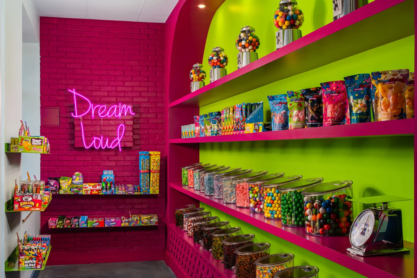 Lime-green shelves of bulk candy jars and packaged sweets against pink brick, beside a pink Dream Loud neon sign.
